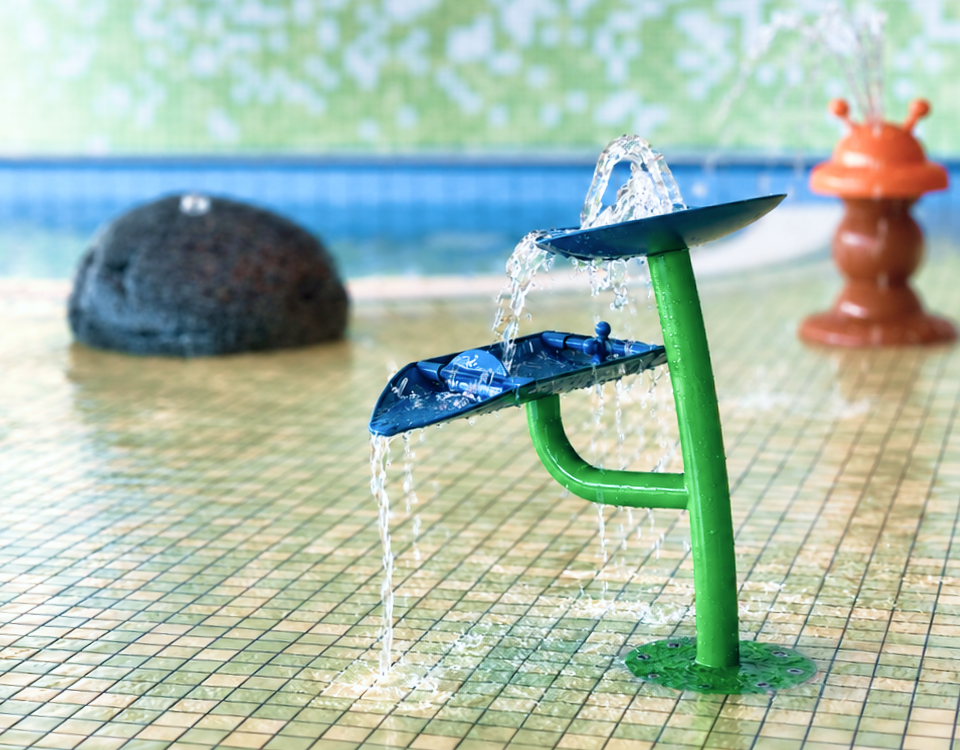 Buntes Wasserspielgerät mit zwei blauen Schalen und grüner Halterung, aus denen Wasser fließt. Im Hintergrund sind weitere Wasserspiele im flachen Kinderbecken zu sehen.